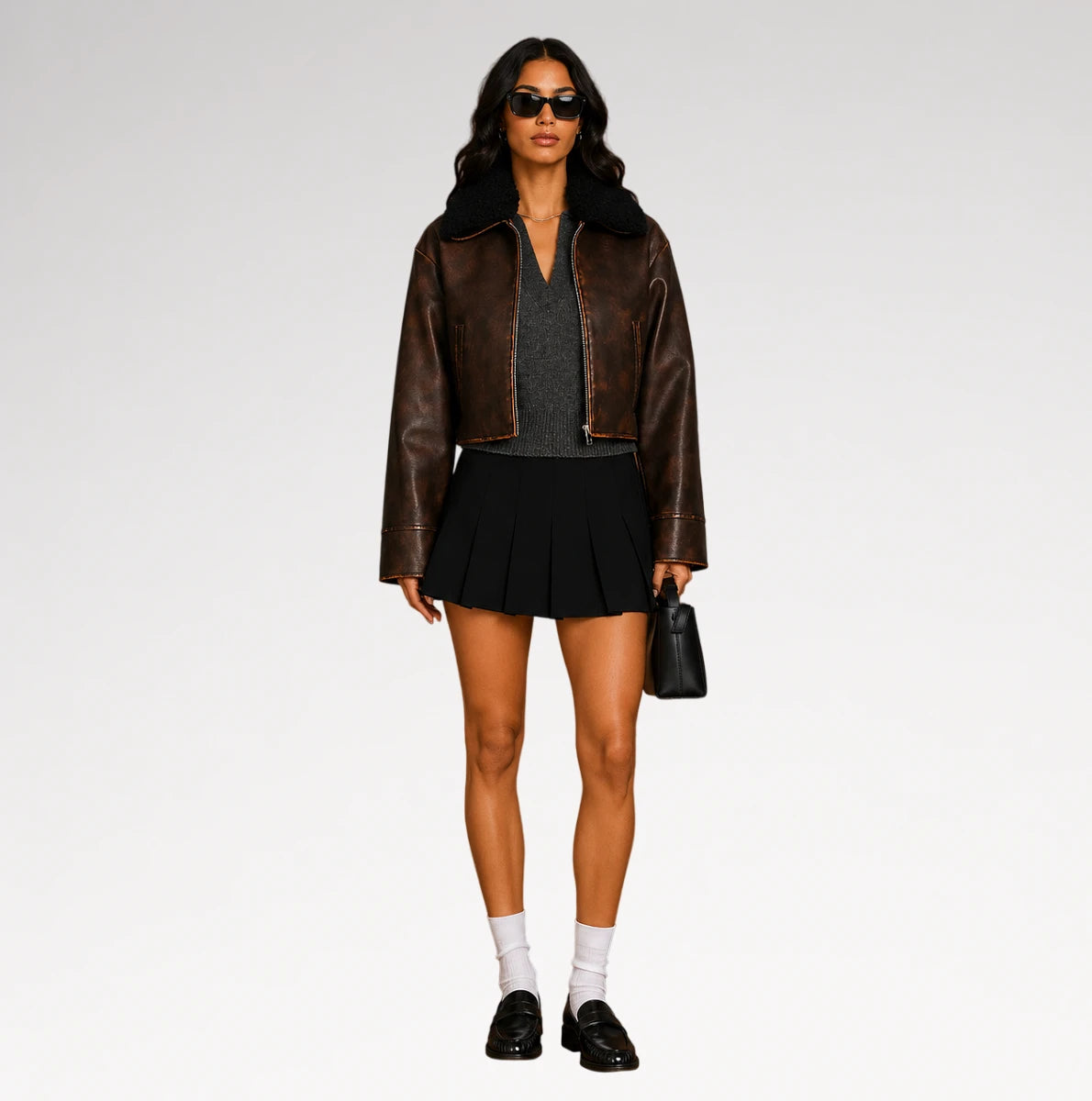 A model stands against a light gray studio background wearing a brown leather jacket with a black shearling-style collar, a dark gray knit top, a black pleated mini skirt, white socks, black loafers, sunglasses, and carrying a small black handbag.
