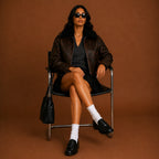 Woman sitting on a chair wearing a brown leather jacket and sunglasses against a brown background