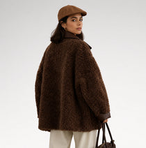 A woman wearing an oversized dark brown teddy-textured coat, a brown flat cap, and cream trousers, holding a dark brown handbag against a light gray studio background.