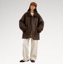 A model stands against a light gray studio background wearing an oversized dark brown jacket with a brown faux-fur collar and cuffs, paired with cream wide-leg trousers, a brown cap, and dark brown shoes.