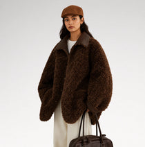 A model wearing an oversized dark brown curly faux-fur jacket with contrasting smooth brown collar and cuffs, styled with a brown flat cap, cream trousers, and a dark brown leather handbag against a light gray studio background.