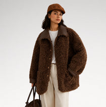 A model wearing a brown textured teddy coat with dark brown collar and pocket trim, paired with cream trousers, a white top, and a brown cap, holding a dark brown handbag against a light gray studio background.