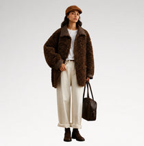 A model stands against a light gray studio background wearing a brown textured teddy-fur jacket with dark brown trim, a white top, cream wide-leg trousers, dark brown ankle boots, a brown flat cap, and carrying a dark brown leather bag.