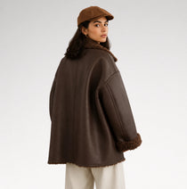 A woman wearing an oversized dark brown faux-shearling coat with a matching brown beret, photographed from the back against a light gray studio background.
