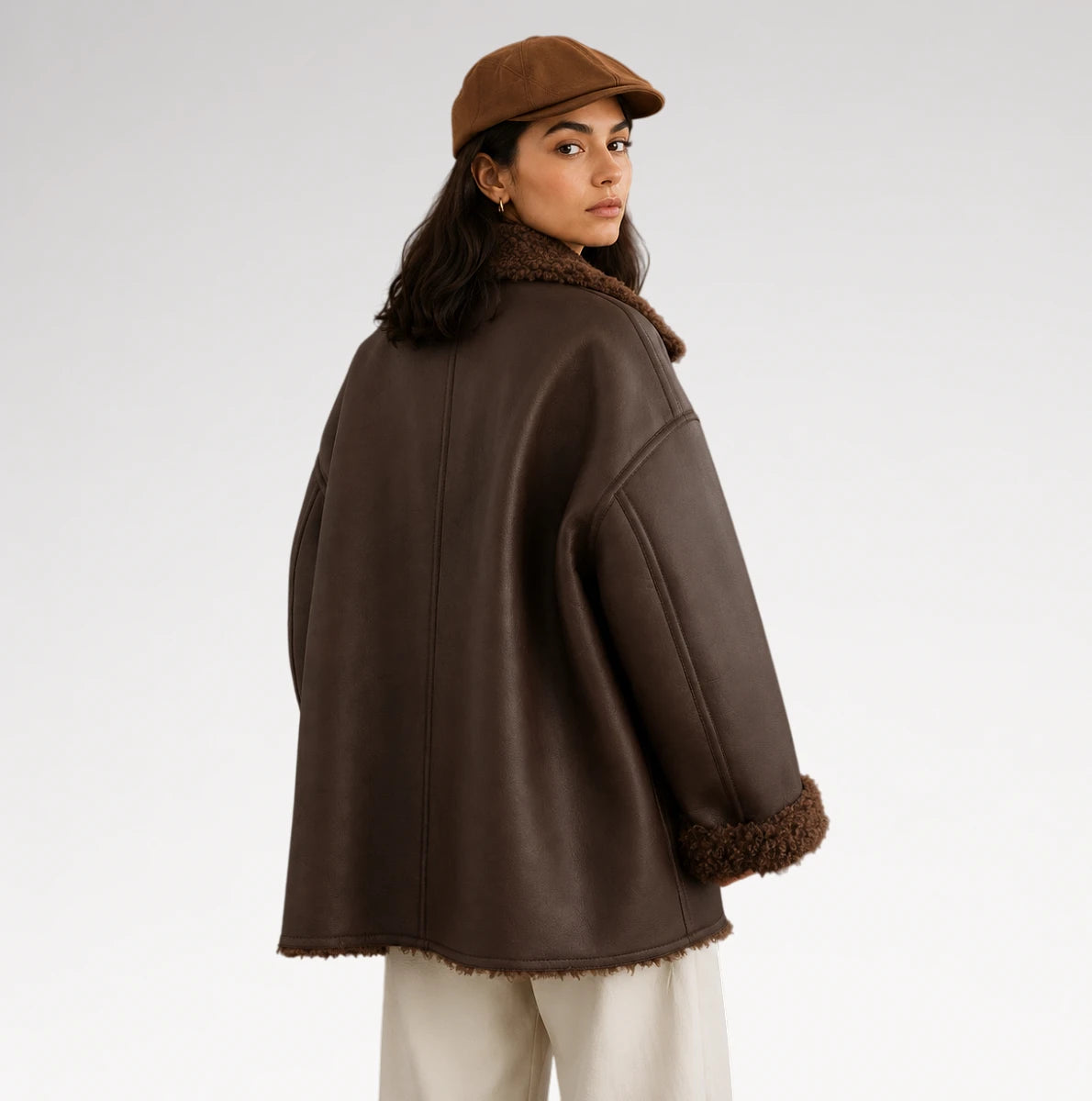 A woman wearing an oversized dark brown faux-shearling coat with a matching brown beret, photographed from the back against a light gray studio background.