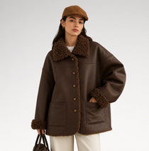 A woman models an oversized dark brown faux-leather shearling jacket with a matching brown flat cap, white pants, and a dark brown handbag against a light gray studio background.