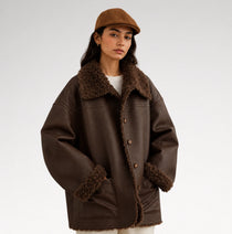 A woman wearing an oversized dark brown faux-leather jacket with a brown shearling-style collar, cuffs, and hem, paired with a matching brown flat cap and a white top, photographed against a light gray studio background.