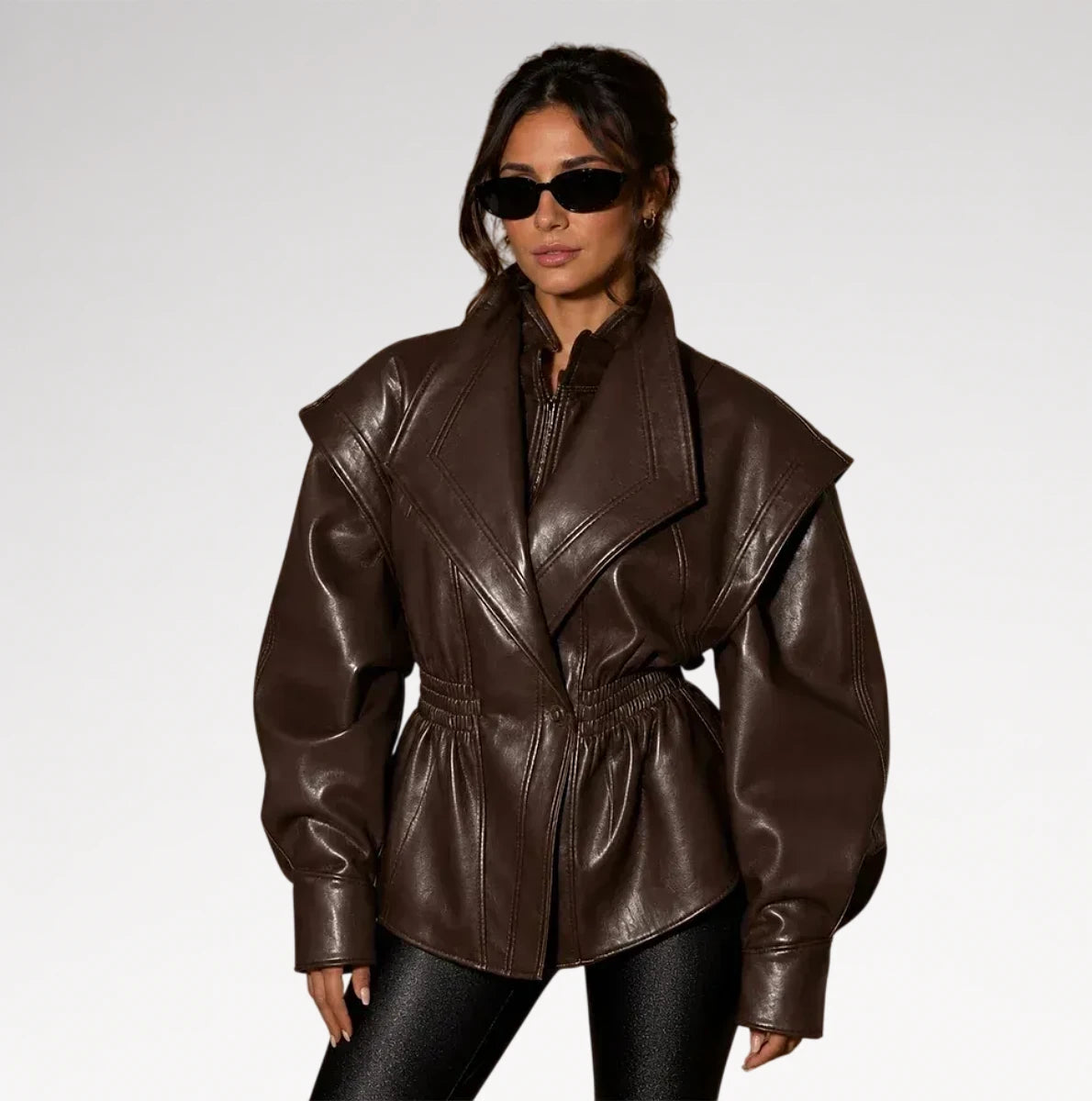 A woman modeling a dark brown faux-leather jacket with dramatic shoulder flaps, a high collar, and a cinched waist, paired with black pants and sunglasses against a light gray studio background. Main image