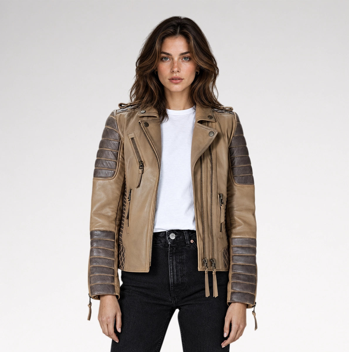 A woman modeling a tan faux-leather moto jacket with dark brown ribbed sleeve panels, a white t-shirt, and black jeans against a light gray studio background.