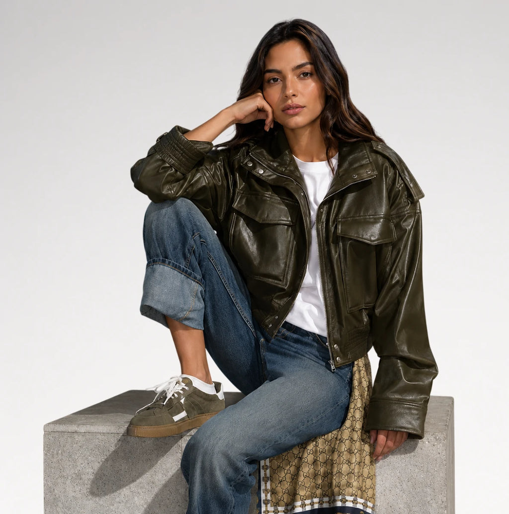 A woman models an olive green faux leather moto jacket, styled with blue jeans, a white t-shirt, and brown sneakers while sitting on a concrete block against a light gray background.