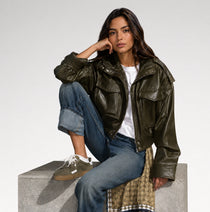 A woman models an olive green faux leather moto jacket, styled with blue jeans, a white t-shirt, and brown sneakers while sitting on a concrete block against a light gray background.