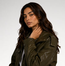 A woman with long, wavy brown hair wearing an olive green faux leather jacket, posing against a light gray studio background.
