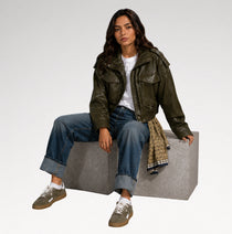 A woman sits on a gray block wearing an olive green leather jacket, blue jeans, white t-shirt, and brown sneakers. A patterned scarf is draped over her leg against a clean white studio background.