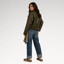 A woman is shown from a rear three-quarter angle wearing an olive green faux-leather moto jacket, blue jeans, and brown shoes, holding a patterned scarf against a plain light gray studio background.
