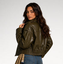 A woman wearing a cropped olive green faux leather jacket, shown from a three-quarters back view against a light gray studio background.