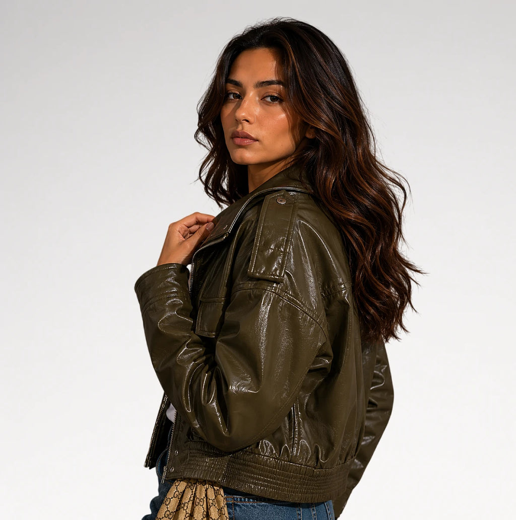 A woman wearing an olive green faux leather jacket, shown from a three-quarters back view against a plain light gray studio background.