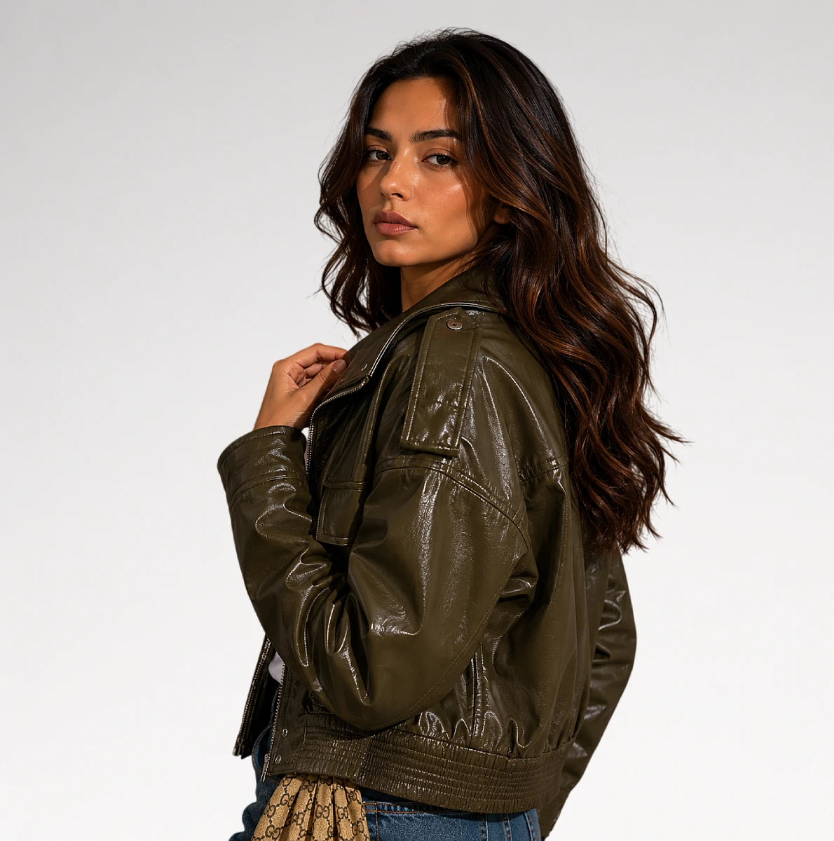 A woman wearing an olive green faux leather jacket, shown from a three-quarters back view against a plain light gray studio background.