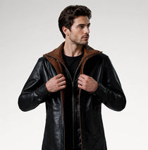 A man wearing a black leather jacket with a brown collar and zipper trim, posing against a light gray studio background.
