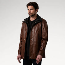 A man wearing a brown leather jacket with a black collar and black shirt, standing against a light gray studio background.