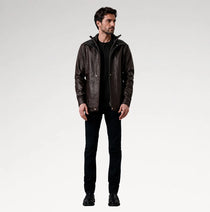 A man modeling a dark brown leather jacket with a high collar, worn over a black shirt and black jeans, standing against a light gray studio background.