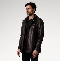 A man wearing a dark brown leather jacket with a high collar over a black shirt, photographed against a light gray studio background.